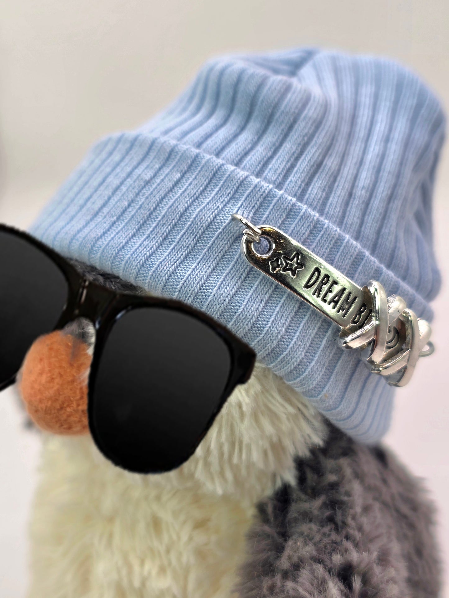 Light blue beanie with 'Dream' charm and sunglasses on a neutral background