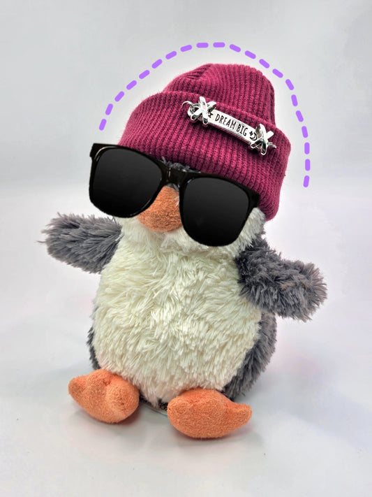 Stuffed penguin wearing a pink beanie with a brooch, black sunglasses, and a gray scarf on a white background.