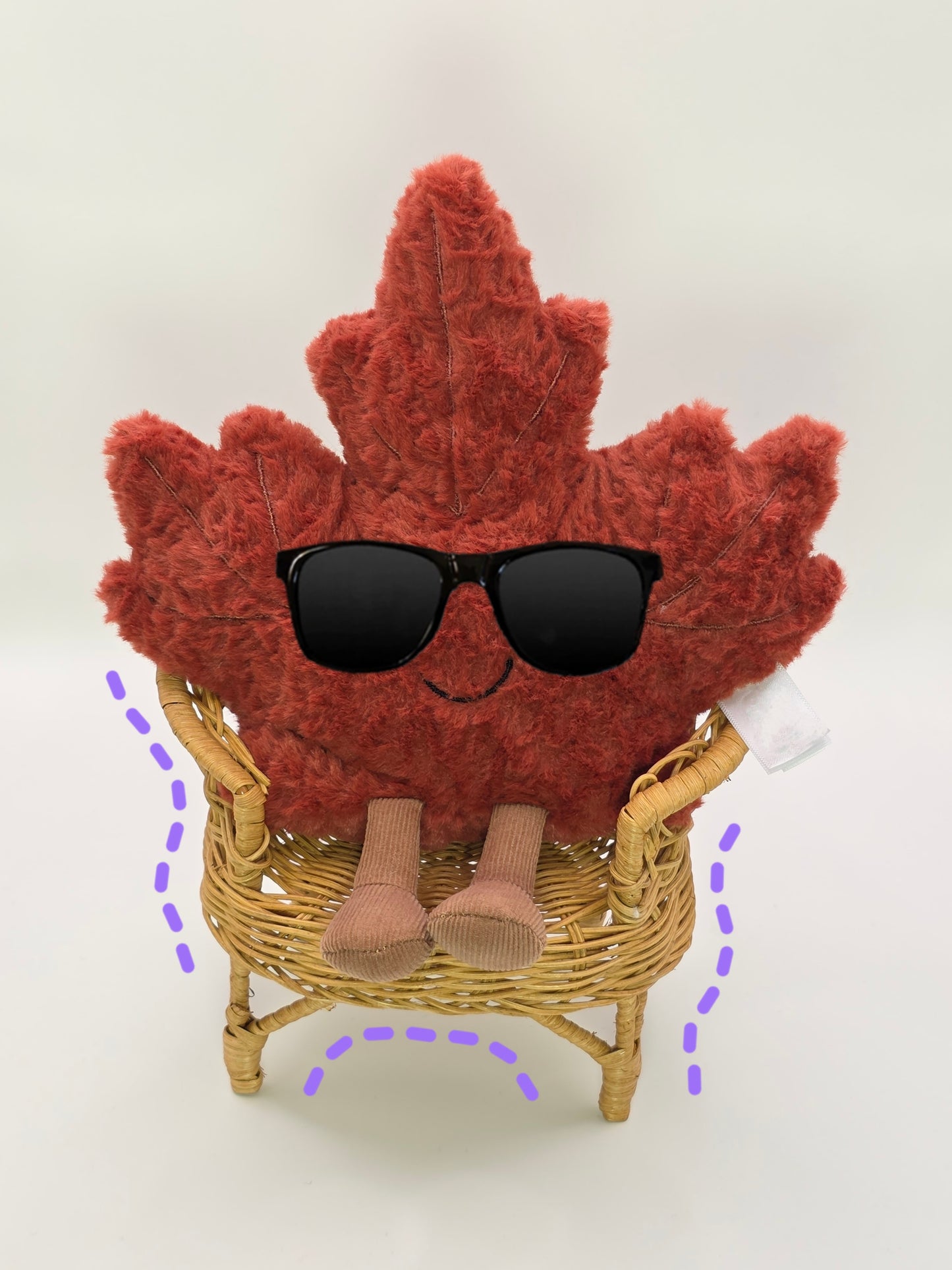 Maple leaf-shaped plush toy with sunglasses sitting on a small wicker chair against a white background
